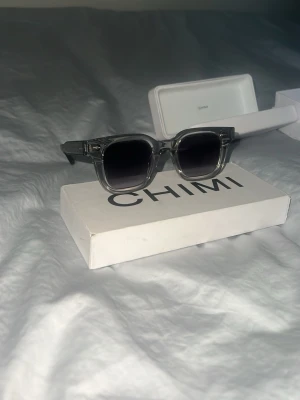 CHIMI solglasögon 04 Grey - Snygga svarta solglasögon från CHIMI med klassisk rektangulär form och silverfärgade detaljer på skalmarna. Glasögonen har en stilren look och passar perfekt för dig som vill ha en lyxig accessoar till din outfit.
