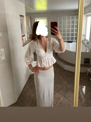 Vit broderad blus med v-ringning - Söt vit blus med broderade detaljer och v-ringning. Modellen är croppad med volang nedtill och har trekvartsärmar med resår vid ärmslut. Perfekt till sommarens outfits och ger en romantisk vibe.