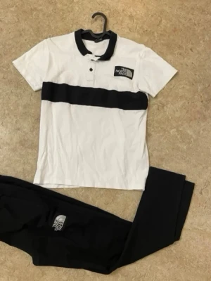 The North Face svartvitt set - Snyggt set från The North Face med en vit pikétröja med svart krage och svart horisontell rand samt matchande svarta byxor. Båda plaggen har tydliga logotyper. Perfekt för en sportig och avslappnad stil.