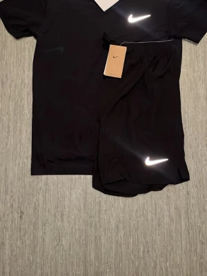 Nike Miller set - Snyggt svart träningsset från Nike med t-shirt och shorts. Båda plaggen har den klassiska vita Nike-loggan framtill. Tillverkade i ett lätt och snabbtorkande material som passar perfekt för träning och sportaktiviteter.