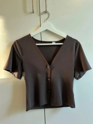 Brun kortärmad topp med knappar - Snygg brun topp med v-ringning och tre dekorativa knappar framtill. Modellen är croppad och har korta ärmar. Perfekt till jeans eller kjol för en enkel men trendig look.
