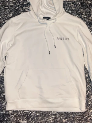 Vit hoodie från Amiri med tryck - Säljer en clean vit hoodie från Amiri med logga tryckt på bröstet och stort AMIRI-tryck på ryggen. Hoodien har huva med dragsko och en stor magficka. Perfekt för dig som gillar streetwear och vill ha något stilrent men ändå fett.