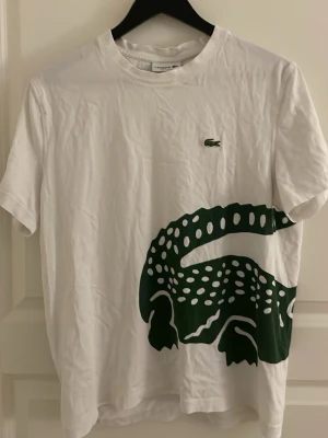 Vit Lacoste t-shirt med stor krokodil - Vit t-shirt från Lacoste med en stor grön krokodiltryck som sträcker sig från framsidan till baksidan. Klassisk rund hals och korta ärmar. Liten Lacoste-logga broderad på bröstet. Perfekt för dig som gillar streetwear och vill sticka ut.
