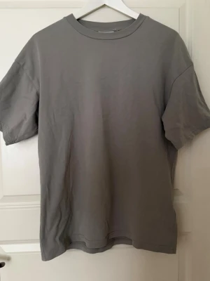 Grå basic t-shirt med rund hals från Weekday - Enkel och stilren grå t-shirt med rund hals och korta ärmar från Weekday. T-shirten har en relaxed passform och är gjord i mjuk bomull som känns skön mot huden. Perfekt till jeans eller shorts för en clean look.