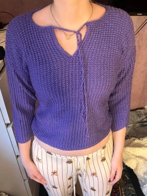 Lila stickad tröja med knytband - Säljer en lila stickad tröja med 3/4-ärmar och grovstickad struktur. Tröjan har en v-ringning med dekorativt knytband framtill och en något croppad passform. Perfekt för dig som gillar färg och vill ha något unikt i garderoben.