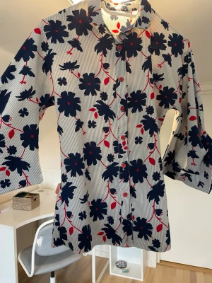 Blommig blus med trekvartsärm - Vit blus med tunna svarta ränder och stora mörkblå blommor samt röda detaljer. Blusen har klassisk krage, knäppning framtill och vida trekvartsärmar. Materialet känns lätt och luftigt, perfekt för dig som gillar färgstarka mönster.