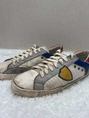 Philippe Model (Retro Edition) - Sneakers från Philippe Model i vitt läder med grå detaljer, blå och röda partier samt en gul sköld på sidan. Klassisk låg modell med vita snören och märkning på plösen. Perfekta för dig som gillar en sportig och färgstark look.