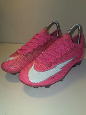 Kylian Mbappe x Mercurial Vapor 13 Elite FG 'Pink Panther' - Snygga fotbollsskor från Nike i en stark rosa färg med vita detaljer och stora Nike-loggor på sidorna. Skorna har snörning och en låg profil, samt ovandel i syntetmaterial som ger bra bollkontroll. Perfekta för dig som vill sticka ut på planen.