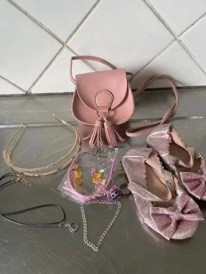 Rosa accessoarset med väska och diadem - Säljer ett gulligt accessoarset med en liten rosa axelväska med tofs, ett par glittriga rosa ballerinaskor med stora rosetter, två diadem (ett guldigt och ett rosa med blommor), samt två halsband och ett armband. Perfekt för att piffa upp din outfit!skorna duger ett tag till 