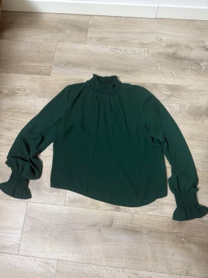 Mörkgrön blus med smockdetaljer - Stilren mörkgrön blus med hög smockad krage och långa ärmar med smockade muddar. Blusen har en lös passform och är tillverkad i ett mjukt, lätt tyg som ger ett elegant fall. Perfekt för dig som gillar trendiga detaljer och en chic look.