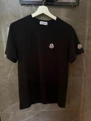 Svart Moncler t-shirt  - Snygg svart t-shirt från Moncler med klassisk logga på bröstet och ärmen. T-shirten har rund hals och korta ärmar, perfekt till en enkel och stilren look. Tillverkad i mjuk bomull för skön känsla hela dagen.