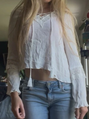 Vit boho blus med spetsdetaljer - Söt vit boho-inspirerad blus med långa ärmar och spetsdetaljer vid ärmslut och halsringning. Blusen har knytband med tofsar framtill och är tillverkad i ett tunt, lätt material. Står att den är i storlek 40 vilket är missvisande då jag tycker den sitter som 36/S. Inga defekter!