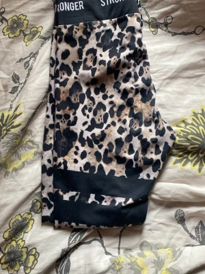 Leopardmönstrade leggings Stronger - Snygga leopardmönstrade leggings från Stronger med bred svart midjeresår där loggan syns. Materialet är stretchigt och bekvämt, perfekt för träning eller chill. Djurmönstret går i beige, brunt och svart för en trendig look.