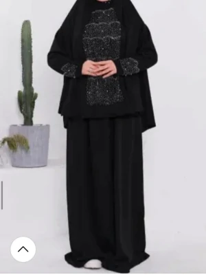 Svart abaya med mönstrad detalj - Säljer en svart abaya med vackra vita mönster på bröstet och ärmsluten. Plagget har en lös passform och täcker hela kroppen. Perfekt för dig som vill ha en modest och stilren look.