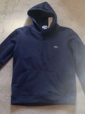 Mörkblå hoodie från Lacoste - Snygg mörkblå hoodie från Lacoste med klassisk krokodillogo på bröstet. Hoodien har huva med dragsko och en stor magficka framtill. Tillverkad i mjukt material som känns skönt mot huden. Perfekt för en avslappnad och stilren look.