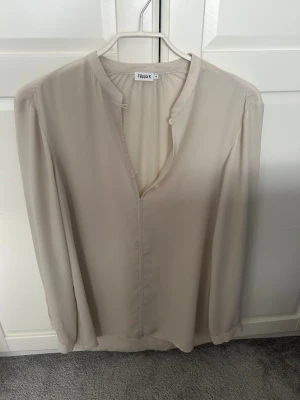 Blus från Filippa K - Stilren vitgråaktig/beige/creme blus från Filippa K med lång ärm och v-ringad hals med liten ståkrage. Tillverkad i ett tunt, lätt och mjukt tyg som ger ett elegant fall. Perfekt för en clean och minimalistisk look.
