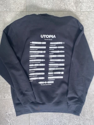 Svart Utopia hoodie Travis Scott - Säljer en svart hoodie från Travis Scott med UTOPIA-tryck och låtlista på ryggen. Hoodien har huva med dragsko, magficka och ribbade muddar. Perfekt för dig som gillar streetwear och vill ha något unikt från artistens merch. Storlek M. Hör av dig vi funderingar👍