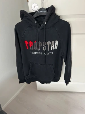 Svart Trapstar hoodie med tryck - Säljer en svart hoodie från Trapstar med stor huva och dragsko. Framtill finns ett färgstarkt Trapstar-tryck i rött, vitt och grått samt texten 'It's A Secret' under. Hoodien har magficka och ribbade muddar vid ärmar och nederkant.