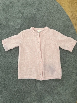 Ljusrosa kofta från Gina Tricot - Söt ljusrosa stickad kofta från Gina Tricot med korta ärmar och rund hals. Koftan har pärlemorknappar framtill och ribbstickade kanter vid hals och ärmslut. Perfekt att slänga över en topp för en chill look.
