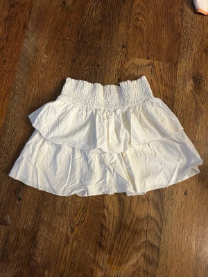 Vit volangkjol med smock - Söt vit kortkjol med bred smock i midjan och dubbla volanger. Kjolen är luftig och har en feminin look, perfekt för varma dagar. Tillverkad i mjukt bomullstyg som känns skönt mot huden. Se bild 2 för fläckar, har ej testat behandla