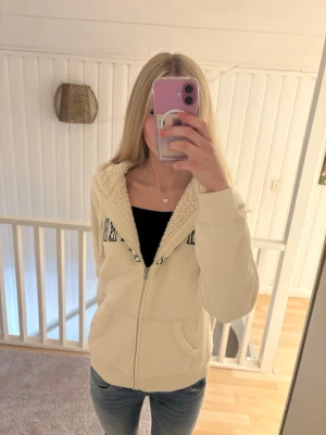 Vit hoodie från Hollister med dragkedja - Säljer en vit hoodie från Hollister med dragkedja och stor huva. Hoodien har mjukt teddyfoder på insidan, två fickor fram och Hollister-logga broderad i mörkblått på bröstet. Perfekt för dig som gillar en mysig och avslappnad stil.