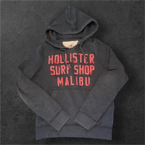 Mörkblå hoodie från Hollister - Mörkblå hoodie från Hollister med stor röd broderad text 'Hollister Surf Shop Malibu' på bröstet. Klassisk huva med snörning och känguruficka framtill. Perfekt för chill dagar och streetstyle. Buda på priset är inte hugget i sten