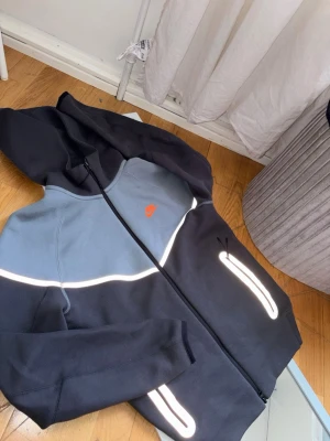 Nike hoodie med reflexdetaljer - Snygg hoodie från Nike i svart och ljusgrått med dragkedja framtill. Hoodien har reflexdetaljer längs bröstet, ärmarna och fickorna samt en liten orange Nike-logga på bröstet. Tillverkad i mjukt material och har en stor huva för extra komfort.