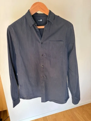 Svart randig skjorta från JC - Klassisk mörkblå skjorta från JC med tunna vita ränder. Skjortan har lång ärm, bröstficka och knappar framtill. Perfekt till jeans eller chinos för en clean look.