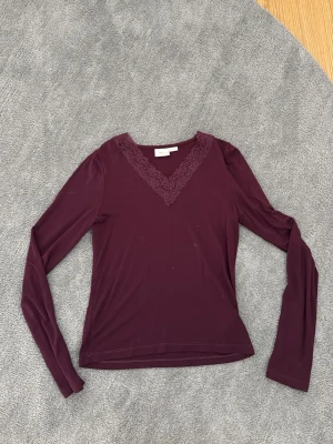 Vinröd långärmad topp med spets - Snygg vinröd långärmad topp med v-ringning och dekorativ spets runt halsen. Perfekt att styla med jeans eller kjol för en trendig look. Materialet känns mjukt och stretchigt.