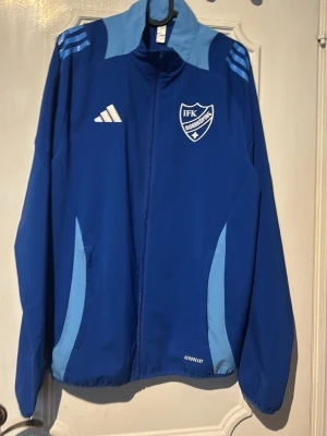 Blå IFK Norrköping träningsjacka Adidas - Snygg blå träningsjacka från Adidas med IFK Norrköping-logga på bröstet. Jackan har ljusblå detaljer på axlar och sidor, dragkedja framtill och lång ärm. Tillverkad i lätt och funktionellt material, perfekt för träning eller uppvärmning.