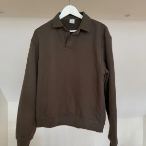 Brun långärmad tröja från Zara - Mörkbrun långärmad tröja från Zara med klassisk krage och knappar vid halsen. Tröjan har en avslappnad passform och är tillverkad i ett mjukt bomullsliknande material. Perfekt för en stilren och enkel look.