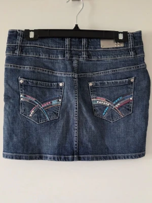 Kort jeanskjol med färgglada detaljer - Snygg kort jeanskjol i blå denim med tre knappar framtill och dekorativa bakfickor med färgglada paljettdetaljer i rosa, blått och silver. Klassisk femficksmodell och normal passform. Perfekt för dig som vill ha en cool och ungdomlig stil.