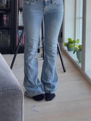 Ljusblå bootcut jeans True Religion - Säljer ett par ljusblå bootcut jeans från True Religion med klassiska kontrastsömmar och detaljerade bakfickor med lock och knappar. Jeansen har låg midja och är tillverkade i mjuk denim med lätt tvättad look. Perfekta för en avslappnad och trendig stil.