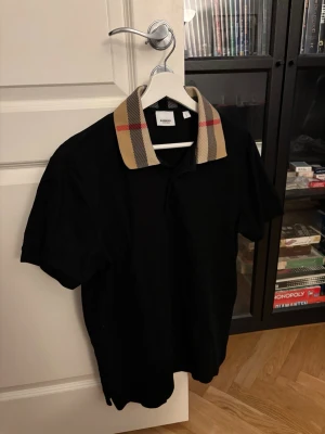 Svart Burberry piké med rutig krage, storlek L  - Burberry Cody Polo Shirt kombinerar en skräddarsydd smal silhuett med varumärkets signatur Check-krage. Tillverkad i bomullspiké, den erbjuder ett raffinerat avslappnat alternativ för vardagsstyling. Viktiga egenskaper: Svart | Bomullspiké | Knappslå | Ribbstickade muddar | Klassisk krage med signatur Check-motiv | Korta ärmar | Mått & passform: Slim fit | Sammansättning & skötsel: 100% bomull -Mycket bra skick
