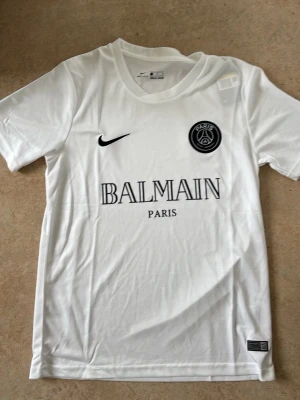 Vit PSG x Balmain t-shirt - Säljer en vit PSG x Balmain t-shirt från Nike med svart logga på bröstet och tryck fram. Klassisk rund hals och korta ärmar. Perfekt för dig som gillar sportmode och vill sticka ut med en unik collab.