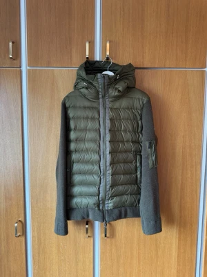 Olivgrön cardigan från Moncler - Säljer en olivgrön pufferjacka från Moncler med stickade ärmar och huva. Jackan har dragkedja framtill, quiltad framsida och en ficka med dragkedja på ärmen.  Storlek M-L passar 180-190. Fattas knap på insidan till fickan men fungerar utan problem. Finns tecken på användning men mycket liv kvar 👏Skriv vid frågor eller bilder | Priset är diskuterbart!