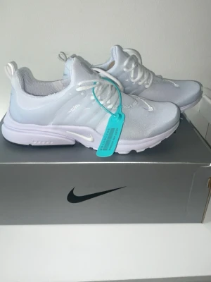 Nike vita air prestos  - Fräscha vita Nike sneakers med mesh-ovandel och vita snören. Skorna har en låg siluett, genomskinliga detaljer på sidorna och en blå zip-tie för extra stil. Swoosh-logga på sidan och tå. Perfekta för dig som gillar clean och sportig look.
