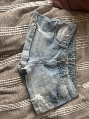 Ljusblå jeansshorts med spetsdetaljer - Säljer ett par ljusblå jeansshorts med snygga spetsapplikationer i vitt och slitna detaljer. Shortsen har klassisk femficksmodell, knappgylf och uppvikta benslut. Perfekta för sommarens alla äventyr!