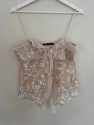 Beige paljettopp från Zara - Supersöt beige linnetopp från Zara med tunna axelband, volangkant upptill och dekorativa paljetter över hela toppen. Toppen har snörning framtill och är perfekt för dig som vill sticka ut lite extra. Lätt och luftig känsla.
