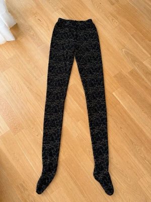 Svarta spetsstrumpbyxor med glitter - Säljer ett par svarta strumpbyxor med vackert spetsmönster och inslag av guldglittrande trådar. De har elastisk midja och är genomskinliga med blommig design. Perfekta för att lyfta vilken outfit som helst med en elegant touch.