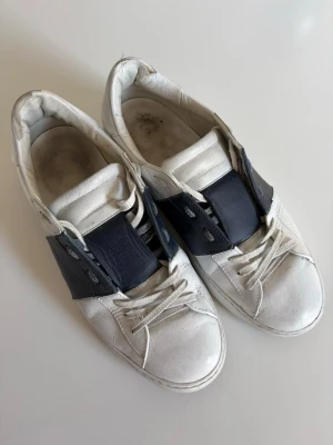 Valentino skor - Säljer ett par vita sneakers med breda marinblå detaljer på sidorna. Skorna har klassisk låg profil, vita snören och rundad tå. Tillverkade i läder för en clean och stilren look. Perfekta för dig som gillar minimalistisk design.