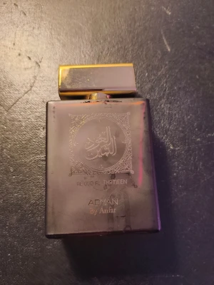 Al Oud Al Thameen parfym Adyan - Elegant parfymflaska i mörklila glas med guldfärgat lock. Flaskan har orientaliskt mönster och texten 'Al Oud Al Thameen' på framsidan. Märket är Adyan by Anfar och designen ger en lyxig och mystisk känsla.