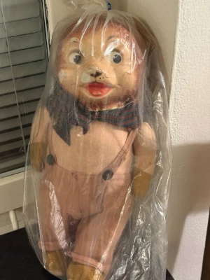 Vintage  - Unik gammal leksaksbjörn med stort huvud, målade ögon och röd mun. Björnen har en beige overall med hängslen och en blå-röd-randig scarf runt halsen. Tillverkad i tyg och troligen papp eller plast i ansiktet. Perfekt för samlare av retroprylar eller dekoration.