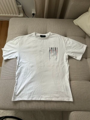 AMIRI t-shirt - Säljer en vit t-shirt med coolt färgstänksmönster och stor logga på ryggen. T-shirten har korta ärmar och normal passform. Perfekt för dig som gillar streetwear och vill sticka ut lite extra.