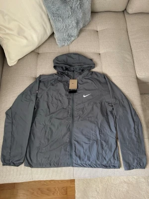 Grå vindjacka från Nike med huva - Snygg grå vindjacka från Nike med huva och dragkedja framtill. Jackan har två sidofickor och en vit Nike-logga på bröstet. Tillverkad i lätt och vindtätt material, perfekt för blåsiga dagar. Enkel och stilren design som passar till det mesta.