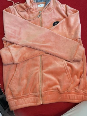 Persikofärgat mjukisset med huva - Mysigt loungewear-set i persikofärgad velour. Setet består av en zip-hoodie med dragsko och fickor samt matchande byxor med resår och dragkedjefickor. Dekorativt H-märke på bröstet. Perfekt för chill dagar hemma.