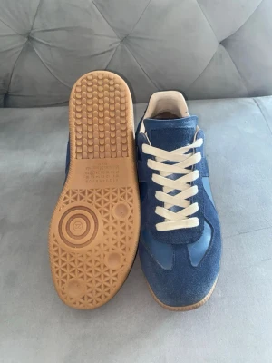 Maison Margiella - Snygga blå sneakers med ovandel i mocka och textil, vita skosnören och klassisk gummisula. Skorna har en låg profil och retroinspirerad design som passar perfekt till jeans eller streetwear. Sulan har ett unikt mönster för extra grepp.