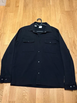 Svart overshirt från Selected Homme - Stilren svart overshirt från Selected Homme med knappar framtill och två bröstfickor med lock. Klassisk krage och långa ärmar med knapp vid ärmslut. Perfekt lager-på-lager-plagg i mjukt material.
