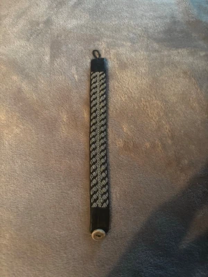 Svart samiskt tennarmband med flätad design - Säljer ett svart samiskt armband med flätad tenntråd och läderdetaljer. Armbandet har ett klassiskt mönster i silverfärgad tenntråd och stängs med en knapp i tenn. Perfekt accessoar för dig som gillar unika och handgjorda smycken.