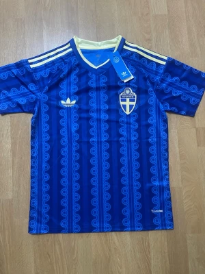  fotbollströja  - Säljer en blå fotbollströja från Adidas med Sveriges landslagsemblem på bröstet. Tröjan har ett mönster i olika blå nyanser, gula detaljer på axlarna och vit krage. Perfekt för dig som vill stötta Sverige på match eller träning.
